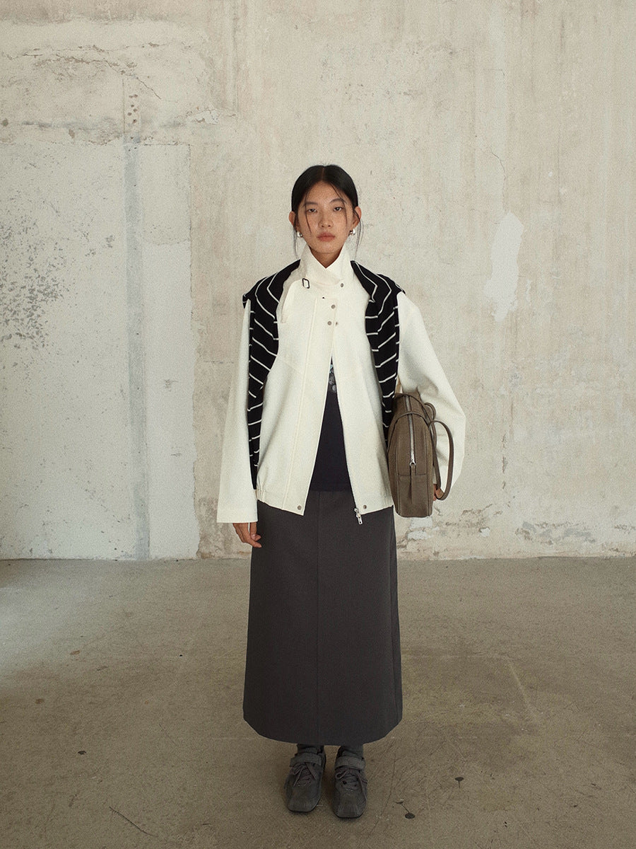 Person wearing a white Stand-up Collar Jacket  with black accents, dark skirt, and carrying a brown bag against a plain wall.